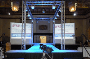 Conference stage with lighting rig above it and two screens either side of the stage