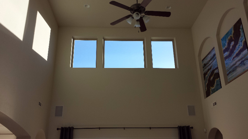 High-ceilinged room with three windows showing blue sky, two paintings, and a ceiling fan.
