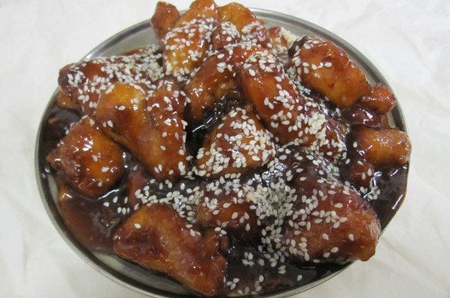 Sesame chicken, covered in sauce, sprinkled with sesame seeds, in a metal bowl.