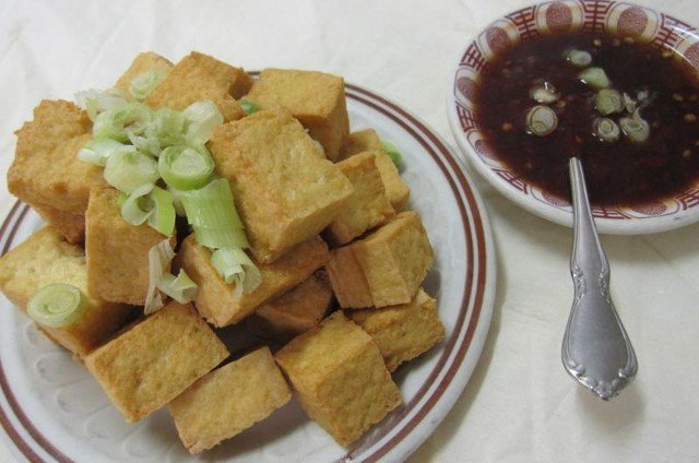 Fried tofu cubes on a plate, garnished with scallions, served with dipping sauce.