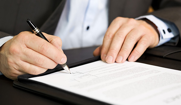 Un uomo con completo seduto alla scrivania, mentre firma un documento con una penna di color nero