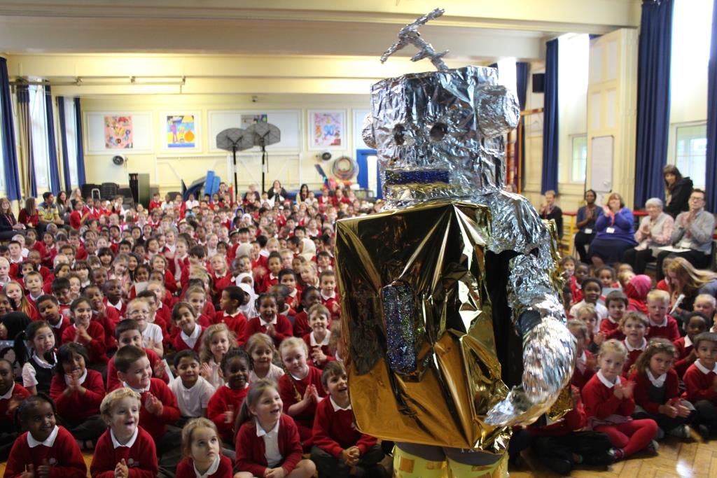 a large group of children are sitting on the floor in a large room . someone is dressed like a robot .