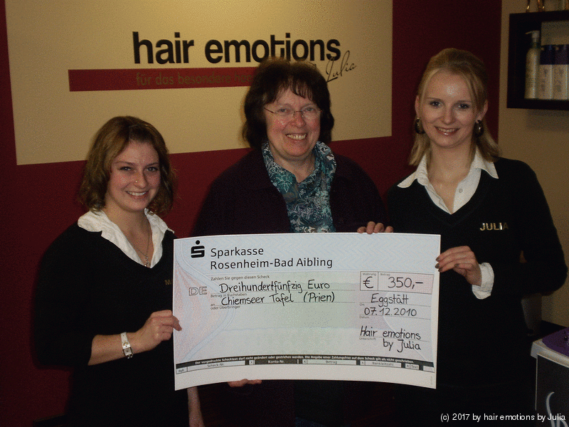 Drei Frauen halten einen Scheck vor einem Schild mit der Aufschrift „Haar-Emotionen“