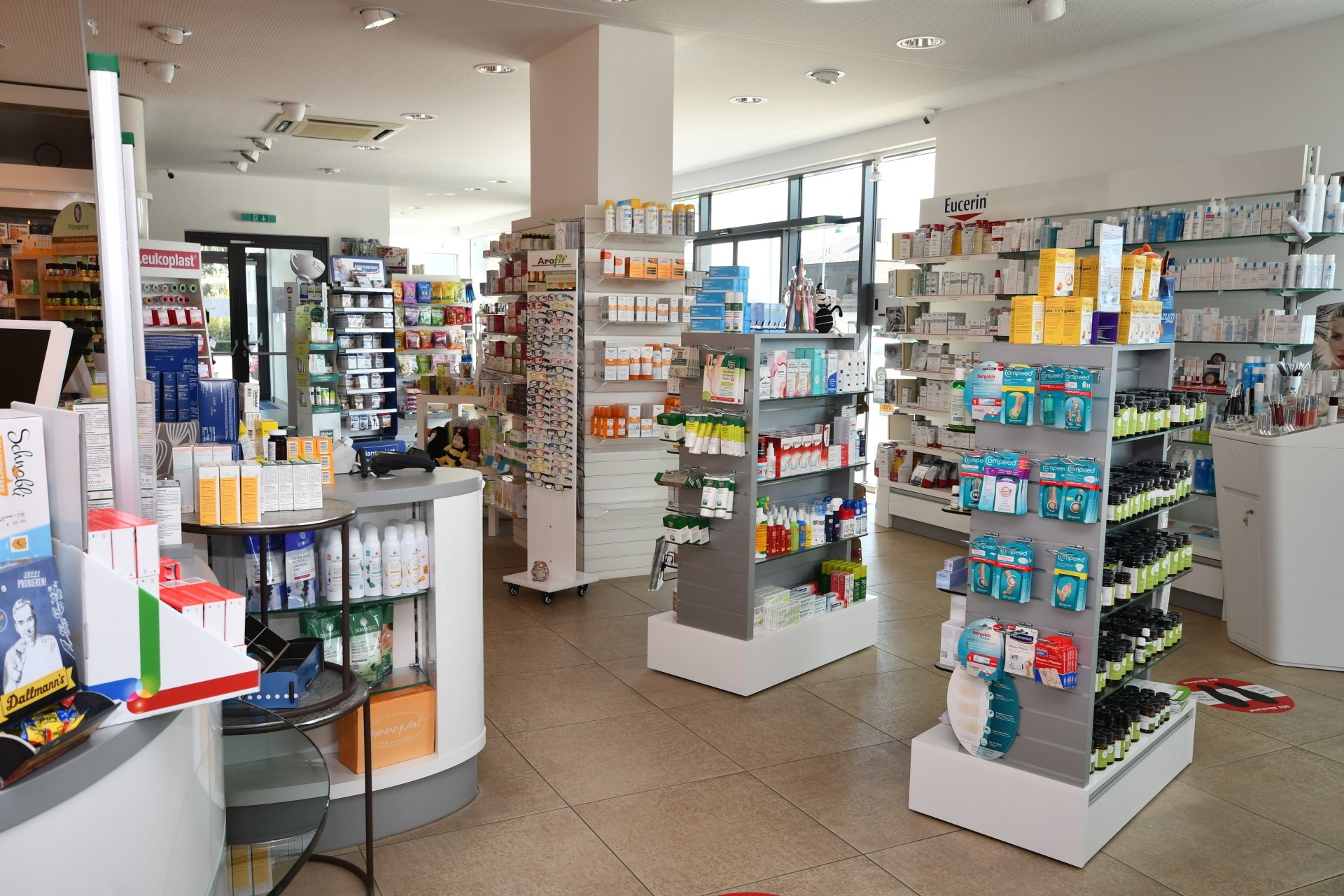 Innenansicht einer gut sortierten Apotheke mit verschiedenen Medikamenten in Regalen und einem Bedienschalter.