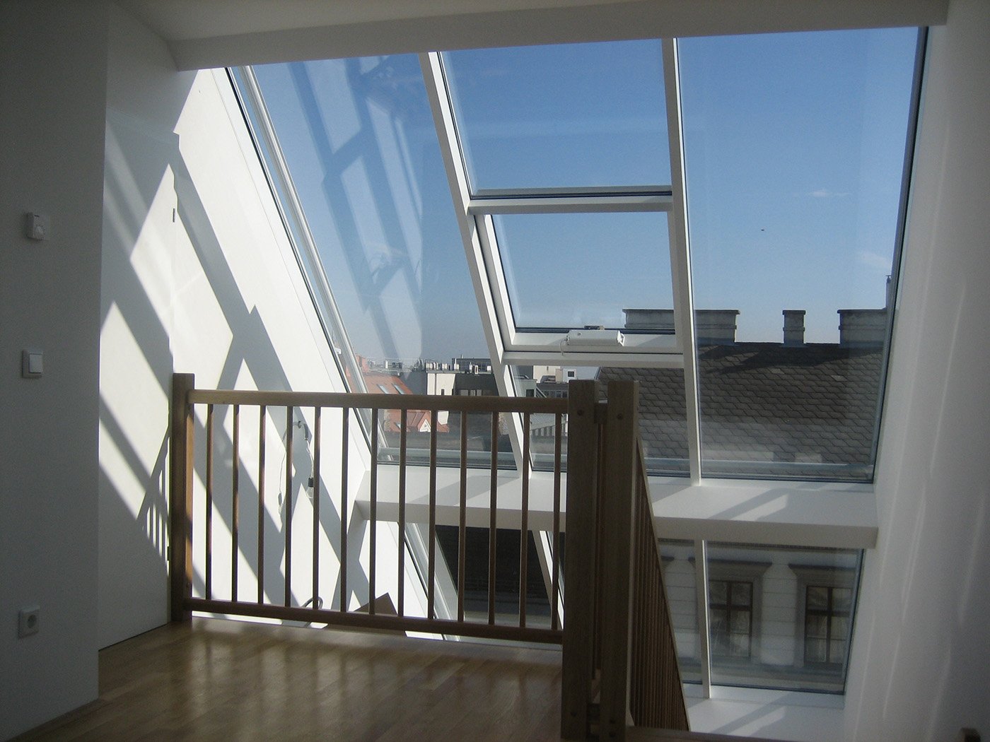 Innenansicht einer Treppe, die zu großen Fenstern mit Blick auf eine Stadtlandschaft und einem Holzgeländer führt.
