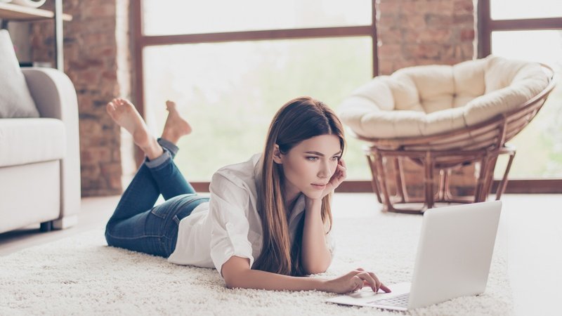 Vrouw ligt op het tapijt en gebruikt een laptop in de woonkamer.