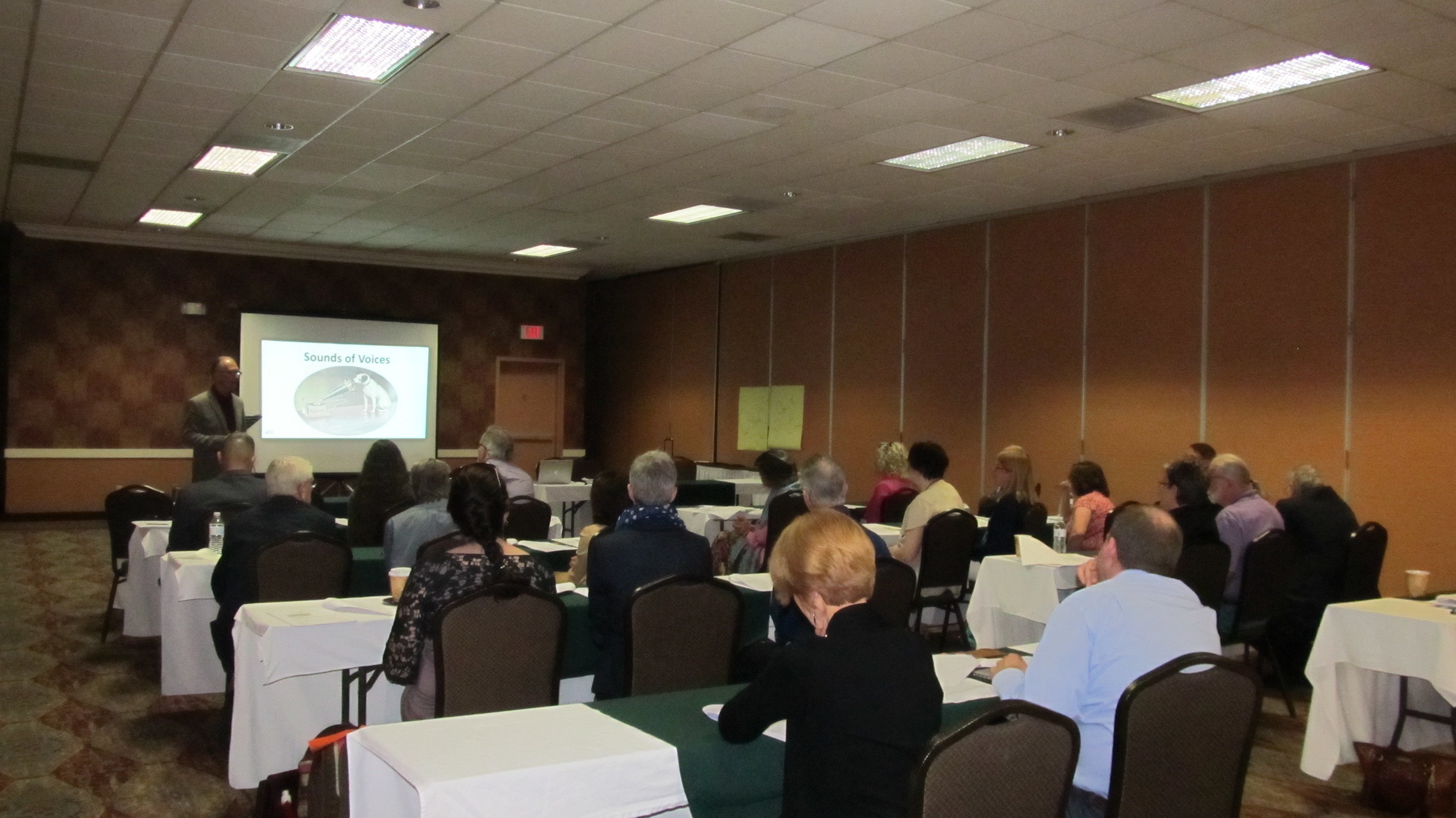 A speaker presents to a seated audience in a conference room with tables and a projection screen.