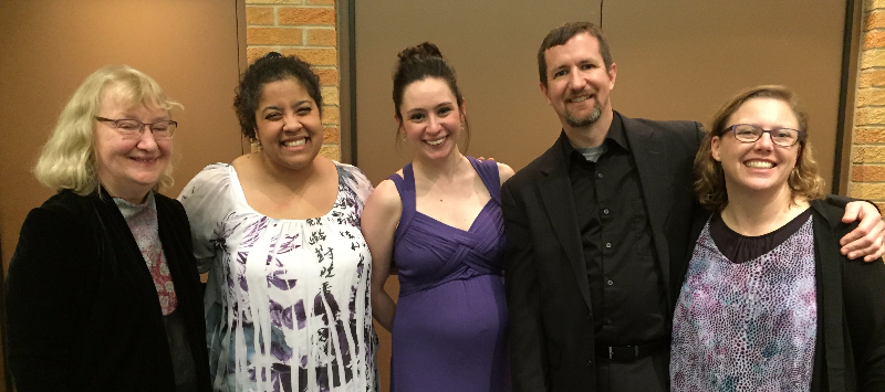 Five people smiling and posing together; the setting appears to be indoors with a neutral background.