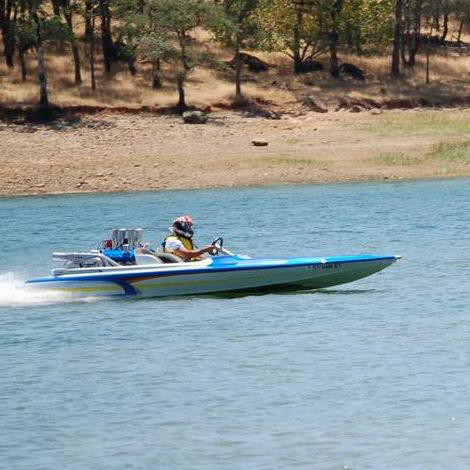 Blue speedboat racing across a lake, leaving a white spray near a dry, wooded shoreline.