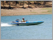 Two people riding a blue motorboat across a lake near a dry, wooded shore