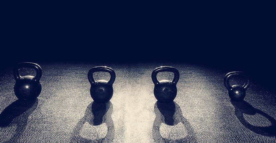 Four kettlebells are lined up in a row on the floor in a dark room.